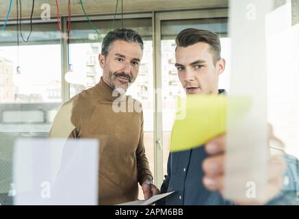 Homme d'affaires et jeune homme d'affaires mûrs travaillant sur des notes adhésives à la vitre au bureau Banque D'Images