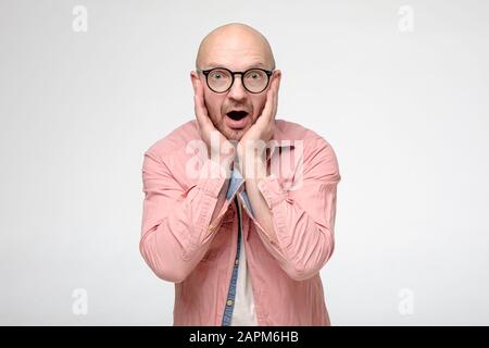Un homme chauve choqué dans les verres tient émotionnellement les mains sur son visage, en surprise ouvre la bouche et regarde dans l'appareil photo avec de grands yeux. Banque D'Images