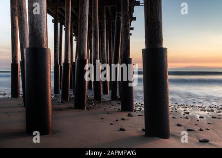 Gros plan des piliers, Ventura Pier, Ventura, Californie au coucher du soleil. Sable et rochers en premier plan; océan soyeux, ciel coloré au-delà. Banque D'Images