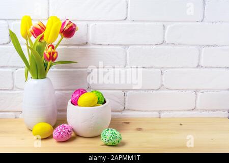 Arrangement rustique de Pâques avec œufs décorés, tulipes rouges et jaunes dans le vase blanc près de mur de briques peintes Banque D'Images