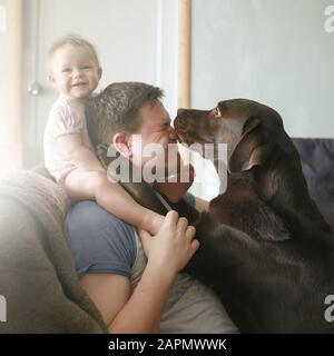 Portrait de famille du jeune père caucasien avec une petite fille souriante mignonne assise sur ses épaules et animal labrador retriever lécher son visage et Banque D'Images