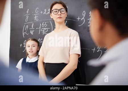 Jeune professeur dans des lunettes se tenant près du tableau noir avec des formules et à l'écoute des enfants de l'école Banque D'Images