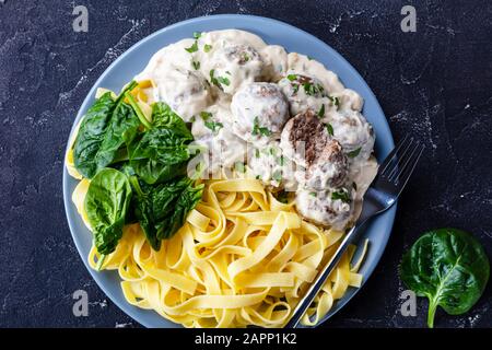 Boulettes de viande suédoise classiques servies avec des nouilles aux œufs et des feuilles d'épinards fraîches sur une assiette à fourche argentée sur une table de cuisine en béton, vue horizontale fro Banque D'Images