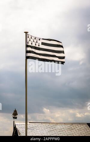 Drapeau de Bretagne, nommé Gwenn ha du en Breton, volant au-dessus d'un toit en ardoise avec une girouette sous forme de bateau à voile contre un ciel de tempête. Banque D'Images