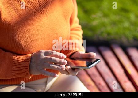 Les mains des femmes tiennent un smartphone et l'utilisent pour discuter. Une femme dans un pull orange est assise sur un banc dans un parc. Banque D'Images