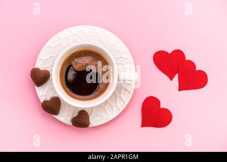 Vue de dessus de la tasse de grains de café avec biscuits au chocolat en forme de coeur et coeurs de papier rouge sur fond rose pastel. Concept de Saint Valentin Banque D'Images