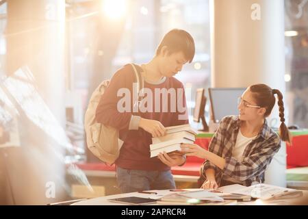 Jeune homme asiatique apportant des livres pour la jeune femme pendant qu'elle travaille à la table Banque D'Images