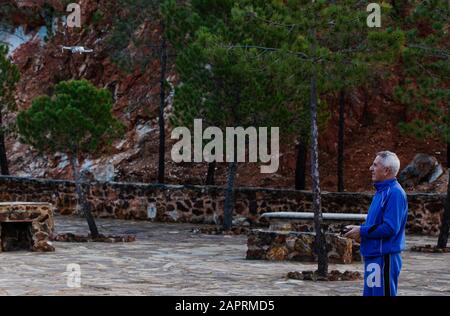 Vue latérale du vieil homme volant la drone dans des vêtements de sport tout en se tenant contre les arbres dans le parc Banque D'Images