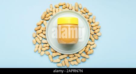 Pâte d'arachide crémeuse dans un pot en verre avec une casquette jaune et des arachides dans le pelure diffusée sur fond bleu pour le petit-déjeuner de cuisine. Concept de nourriture végétalienne Banque D'Images