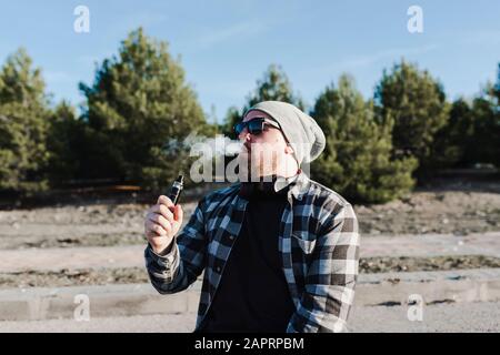 Jeune homme assis sur une longue planche à fumer avec une cigarette électronique Banque D'Images