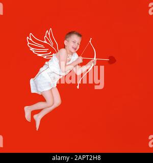 Heureux sourire bébé cupid dans les ailes d'ange de costume, arc et la flèche de coeur isolé sur fond de studio rouge. CopySpace pour le texte. Concept de Saint Valentin Banque D'Images
