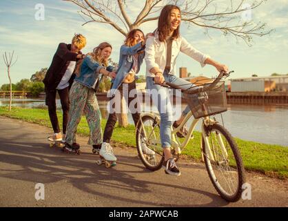 Quatre adolescents qui s'amusent à l'extérieur, à côté d'une rivière, avec patins à roulettes et vélo Banque D'Images