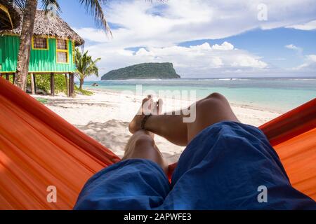 Paire de jambes reposant sur un hamac orange sur une plage de sable blanc Banque D'Images