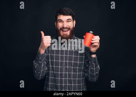 Un jeune homme souriant barbu tient des pouces vers le haut et une tasse se prend une boisson chaude près du fond noir. Banque D'Images