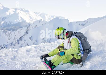 le freerider de snowboarder est assis sur une pente et fastens son snowboard tout en regardant les montagnes. Russie Sotchi Rosa Khutor. Banque D'Images