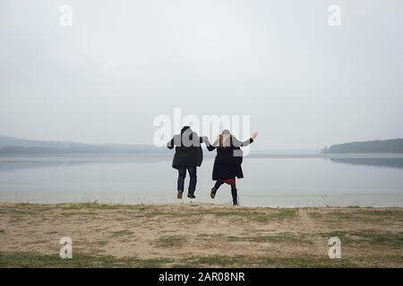 Couple sautant tout en tenant les mains sur une rive de lac Banque D'Images