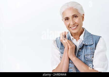 Sensibilité, vieillissement et concept de cosmologie. Femme senior moderne, mignonne et heureuse, souriante, avec ses cheveux gris peignés, ses mains clasues et son appareil photo Banque D'Images