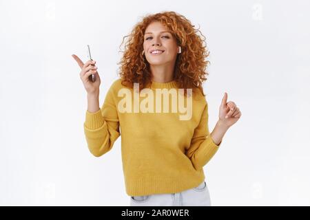 Une jeune fille joyeuse et moderne qui dansait avec une jolie tête rouge sur la chanson préférée, porte des écouteurs sans fil, pointe les doigts et se déplace vers la musique rythmique, tenez-vous Banque D'Images