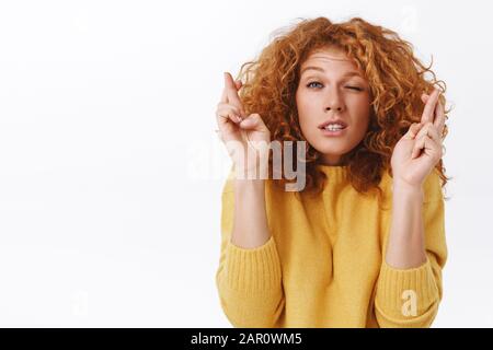 Portrait gros plan une jolie femme mausseuse à tête rouge, pleine d'espoir et inquiète dans un pull jaune, qui s'attonne et grimace aux yeux fermés et aux dents trempées, traverse Banque D'Images