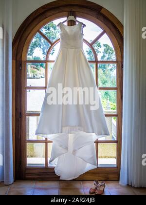 Robe de mariage simple suspendue d'une grande fenêtre avec un jardin sous la lumière du soleil sur l'arrière-plan Banque D'Images