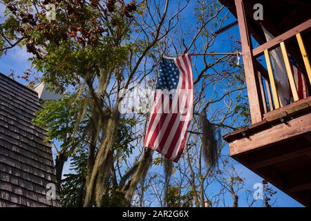 Le drapeau américain vole haut d'un balcon avec un arbre avec des mousses espagnoles suspendues, un ciel bleu et une maison avec des bardeaux de cèdre en arrière-plan. Banque D'Images