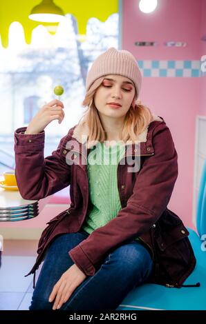 Jolie petite jeune femme vêtue d'un pull, d'une casquette et d'une veste assis dans un café avec des bonbons à la lollipop Banque D'Images