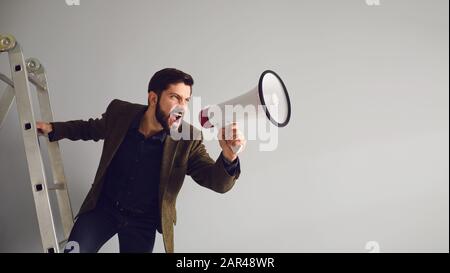 Un homme d'affaires barbu dans les escaliers avec un mégaphone dans les mains se déchèrent sur un fond gris. Banque D'Images