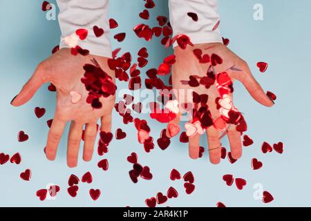 Les mains de la femme jettent des confettis pailletés en forme de coeur rouge sur fond bleu Banque D'Images