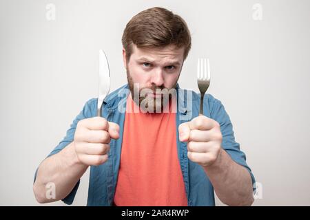 Stern, homme barbu affamé tient un couteau avec une fourchette dans ses mains et semble angry. Concept de régime alimentaire, de crise ou d'économie d'argent. Banque D'Images