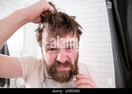 L'homme surcultivé, avec une coiffure et une barbe chignées, regarde dans sa réflexion dans le miroir et est insatisfait de son apparence. Banque D'Images