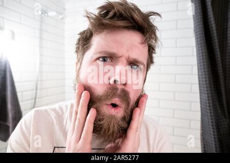 L'homme surcultivé, avec une coiffure et une barbe chiches, se voit se refléter dans le miroir, il est choqué par son apparence. Banque D'Images