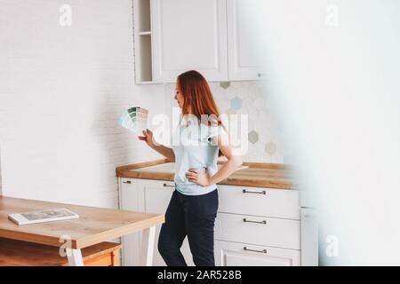 Jeune femme designer avec de longs cheveux rouges tenant des échantillons de couleur dans la cuisine lumineuse à la maison Banque D'Images