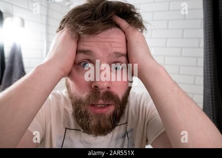 L'homme voit sa réflexion dans le miroir, il est choqué par son apparence chatousseuse et dans les clenches d'horreur la tête dans les mains. Banque D'Images