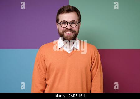 Très mignon homme barbu caucasien dans des verres ronds, dans un pull orange regarde l'appareil photo et sourires. Banque D'Images