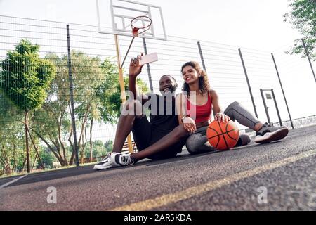 Activité En Plein Air. Couple africain assis sur un terrain de basket-ball prenant selfie sur smartphone souriant heureux Banque D'Images