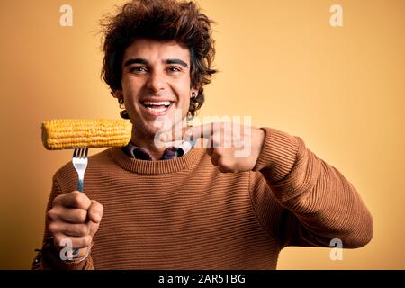 Jeune homme de handsome tenant la fourche avec le maïs rafles debout sur fond jaune isolé très heureux pointant avec la main et le doigt Banque D'Images