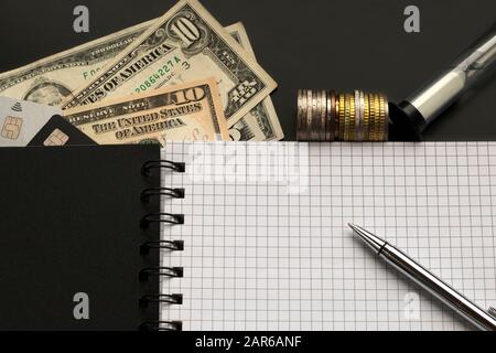 Page vierge avec stylo, dollars, pièces de monnaie, cartes de crédit, verre de sable sur fond noir. Planification, concept de budgétisation Banque D'Images