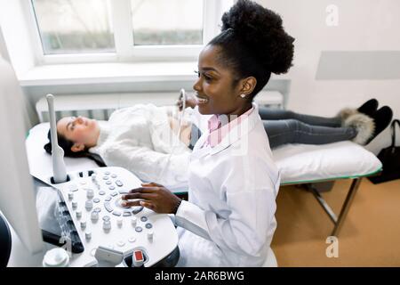 Le médecin généraliste africain souriant vérifie l'abdomen féminin à l'aide d'un équipement ultra-sonore moderne Banque D'Images