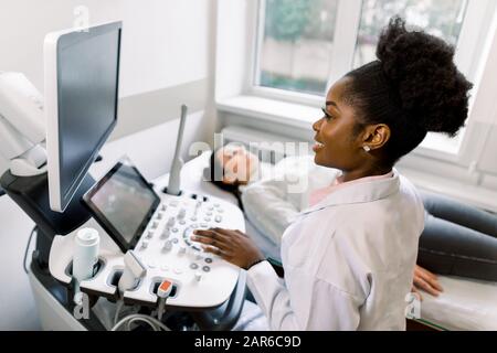 Le jeune médecin africain féminin utilisant une machine à ultrasons en clinique, fait l'échographie abdominale pour la femme enceinte. Scanner À Ultrasons. Sonographie. Banque D'Images
