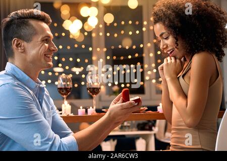Date Romantique. Jeune couple multiethnique à la maison ayant le dîner homme debout sur le genou proposant à la femme avec proposition anneau souriant surpris Banque D'Images