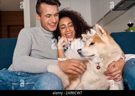 Date Romantique. Jeune couple multiethnique à la maison assis sur un canapé jouant avec le chien riant gai Banque D'Images