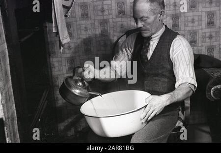 Années 1950, historique, faire de l'hydromel, un homme âgé assis près d'un vieux four, versant de l'eau chauffée d'une bouilloire en métal dans un bol en émail, pour faire l'ancienne boisson alcoolisée hydromel, également connu sous le nom de vin de miel, Angleterre, Royaume-Uni. Banque D'Images