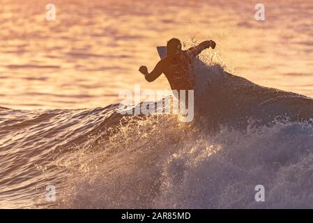 Le candidat surfeur en maillot de compétition est silhouetté contre l'eau réfléchissante de l'océan pendant l'heure d'or tout en réalisant un tour amusant sur une vague. Banque D'Images