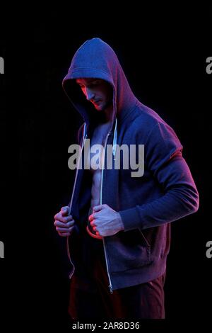 Jeune athlète masculin dans un sweat à capuche bleu foncé non zippé debout dans l'obscurité Banque D'Images