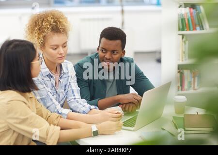 Portrait en grand angle d'un groupe multi-ethnique d'étudiants utilisant ensemble un ordinateur portable tout en travaillant sur un projet dans une bibliothèque, espace de copie Banque D'Images