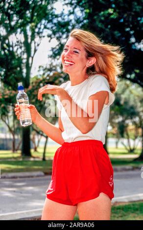 Un jeune jogging féminin blond pour adulte prend une pause pour prendre une boisson rafraîchissante à partir d'une bouteille d'eau dans un MODÈLE de long Beach, CA, City Park Banque D'Images