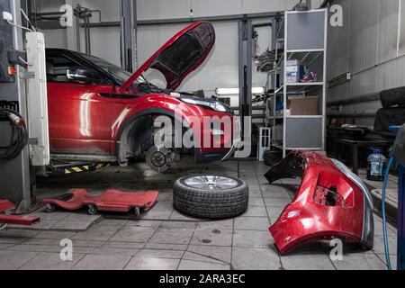 Véhicule d'occasion rouge avec capot ouvert relevé sur un pont élévateur pour la réparation du châssis et du moteur avec roues déposées et pare-chocs cassé dans un atelier de réparation de véhicule. A Banque D'Images