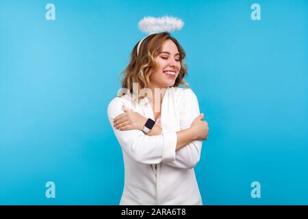 Portrait d'une belle femme angélique égoïste avec halo sur ses cheveux ondulés et en vêtements blancs s'embrassant, debout avec les yeux fermés du plaisir Banque D'Images