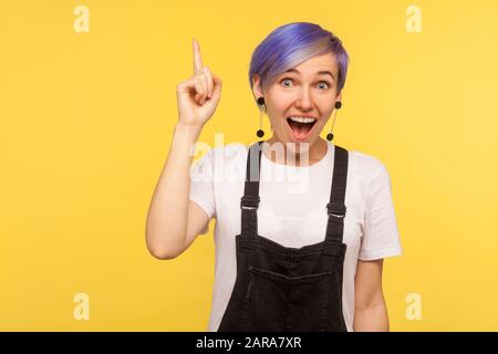 Eureka ! Portrait d'une fille hipster enthousiaste avec des cheveux courts violets dans des salopettes en denim pointant vers le haut, ayant une grande solution d'idée, créatif intelligent Banque D'Images