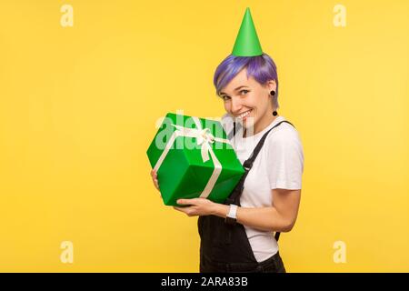 Cadeau d'anniversaire. Portrait d'une jolie fille hipster avec des cheveux courts violets portant un drôle de cône de fête et des salopettes, tenant la boîte enveloppée, heureux avec Banque D'Images